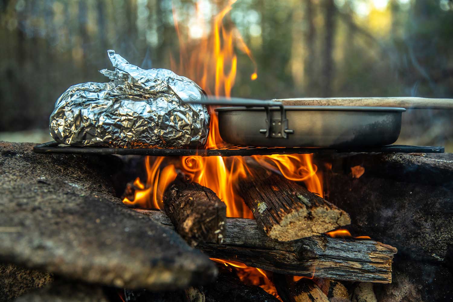Food wrapped in foil and a pan cooking over a campfire.