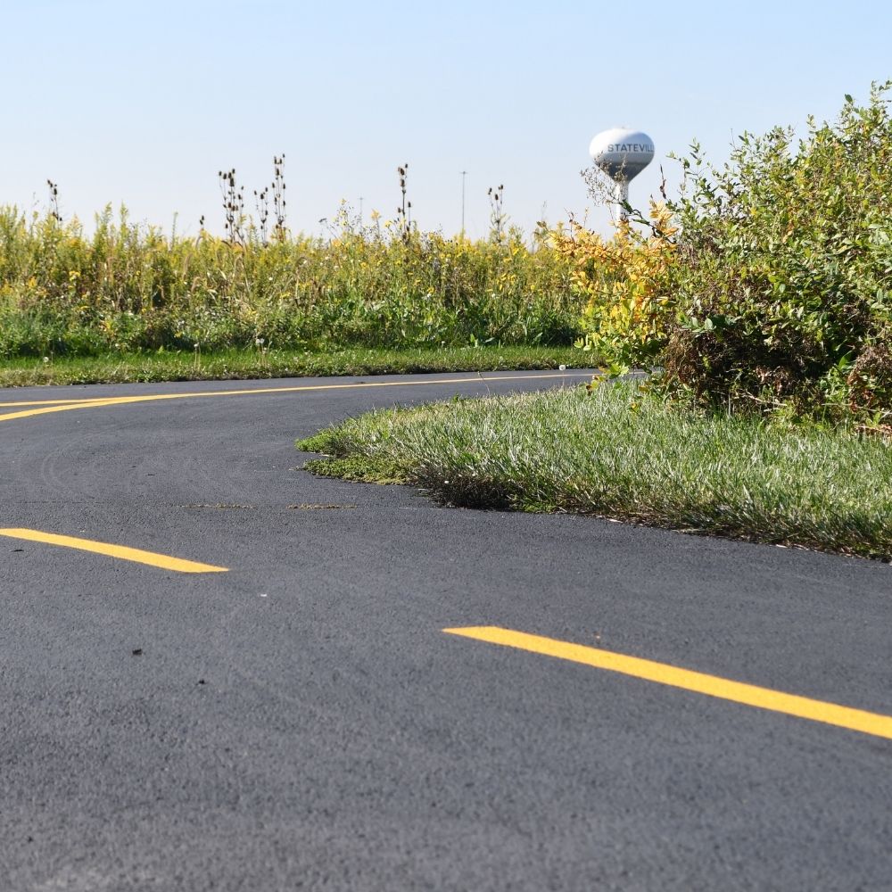 A smoothly paved trail curves through a grassy prairie landscape under a clear blue sky. Tall wild grasses and shrubs line the path.