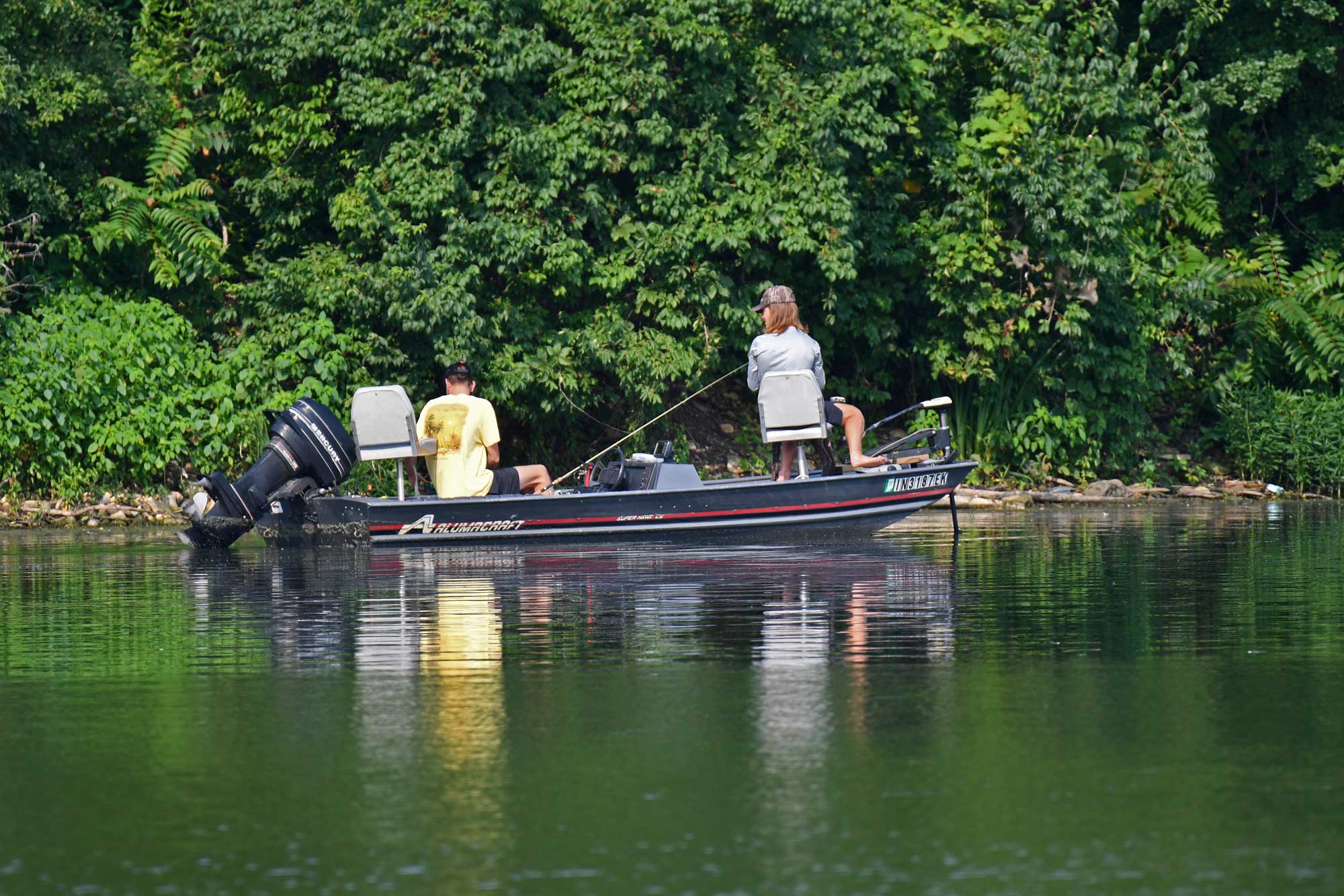 Two people sitting in a boat while fishing with trees in the background.