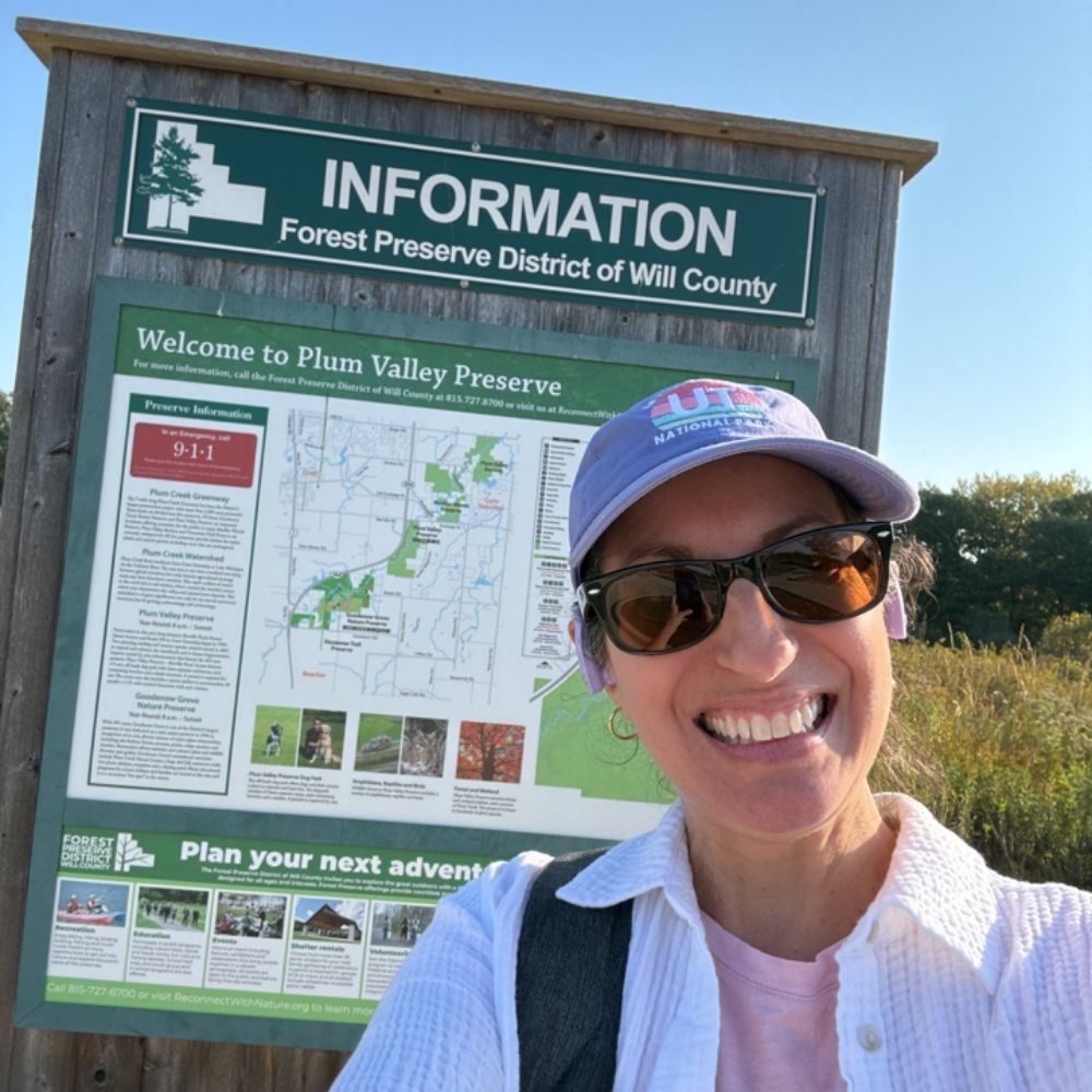 A smiling person wearing sunglasses and a light-colored cap takes a selfie in front of an informational sign for Plum Valley Preserve.
