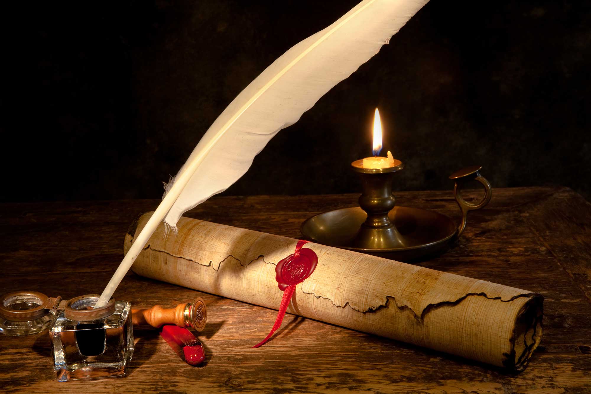 An old-style quill pen next to a paper scroll on a table lit by candle. 