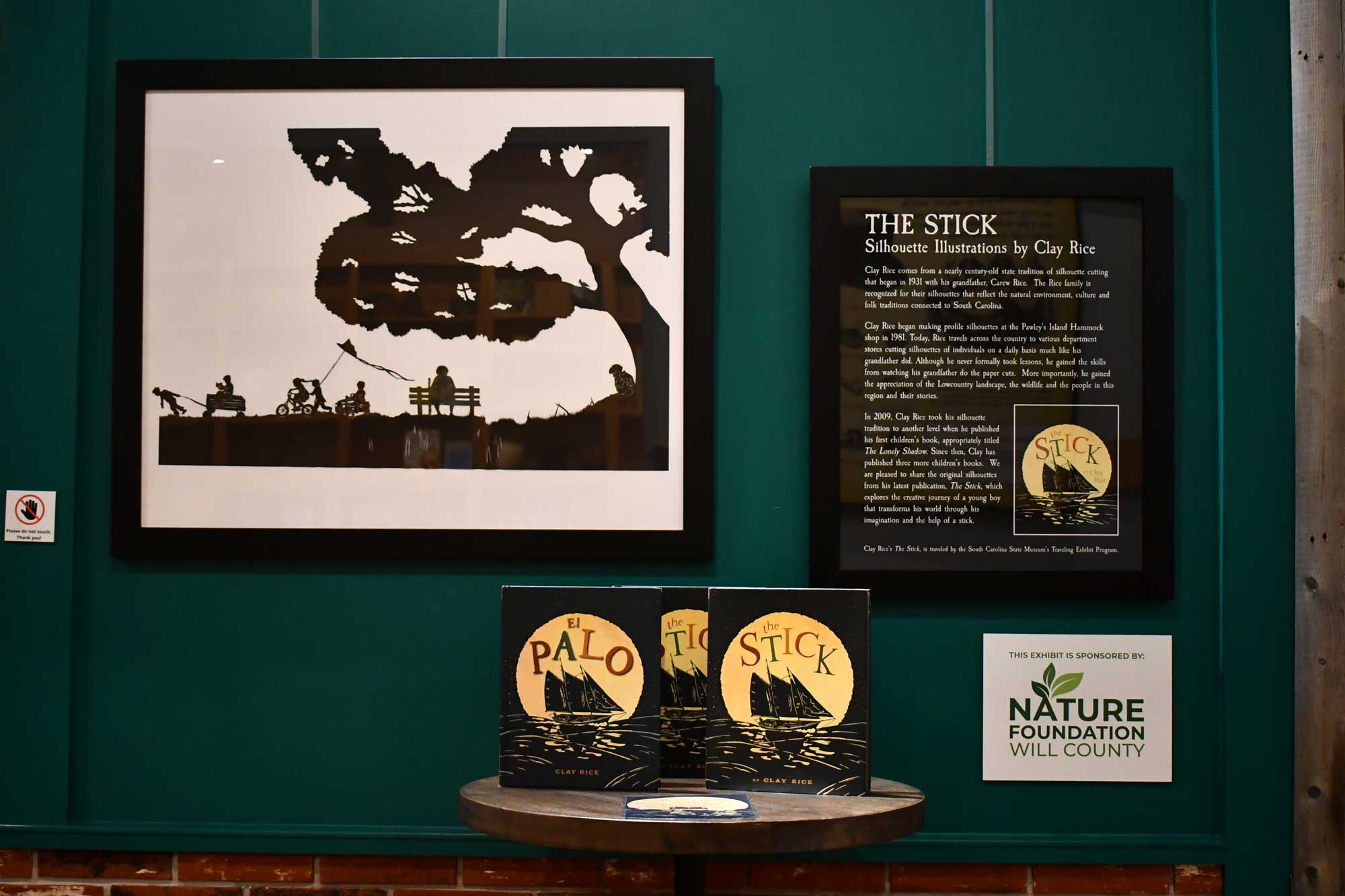 A framed black-and-white illustration of people under a tree. Next to the is a framed synopsis of the book "The Stick" with copies of the book arranged in a table underneath.