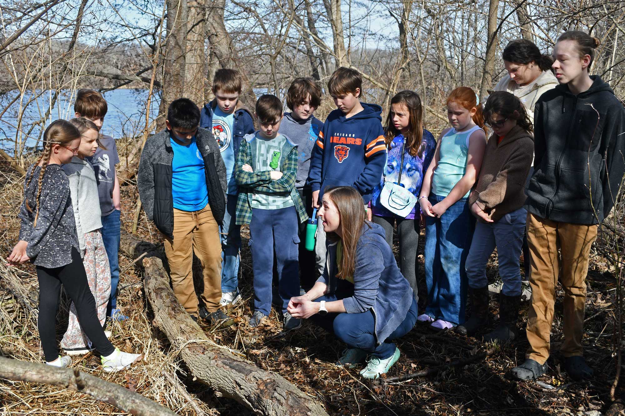 A group of children standing around an adult crouched on the ground and talking to the group.