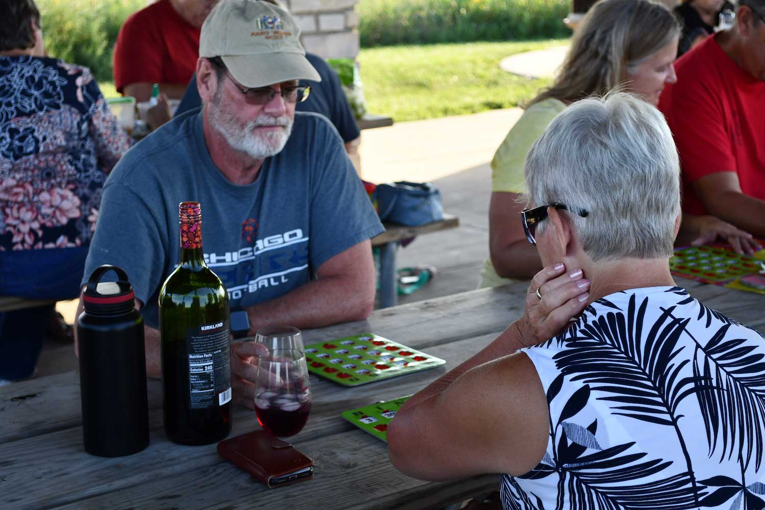 Two people sitting at a wooden picnic table with bingo cards. A bottle of wine and wine glasses are on the table, and more people are in the background.