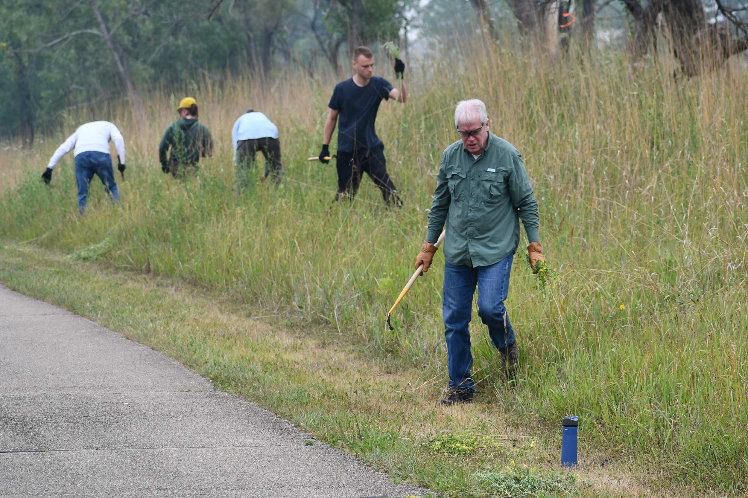 A group of people working in a grassy area next to a paved trail. Some are holding tools and others are bent over at the waist.