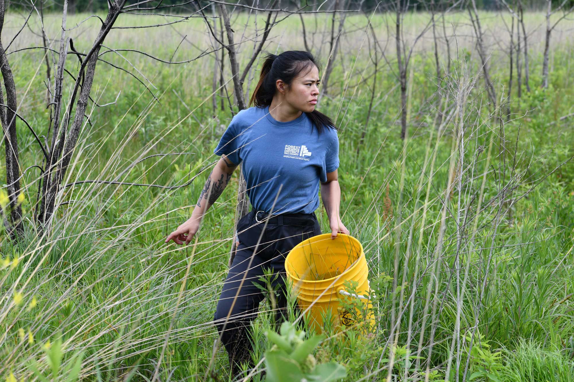 A person holding a yellow 5-gallon bucket and looking backward while standing in a prairie. 