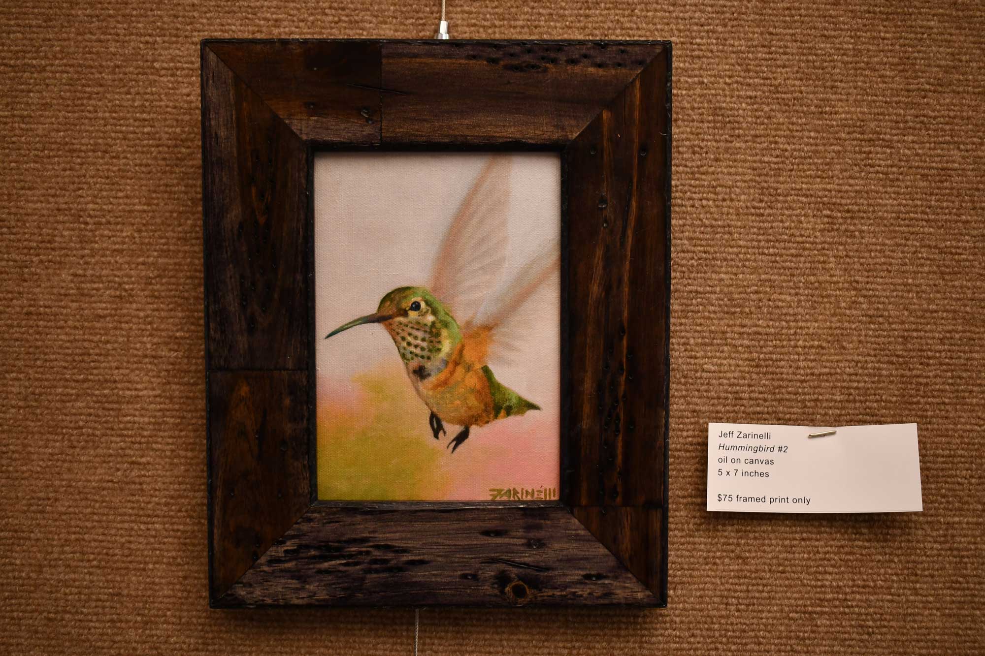 A painting of a hummingbird in flight in a wooden frame hanging on a fabric wall.