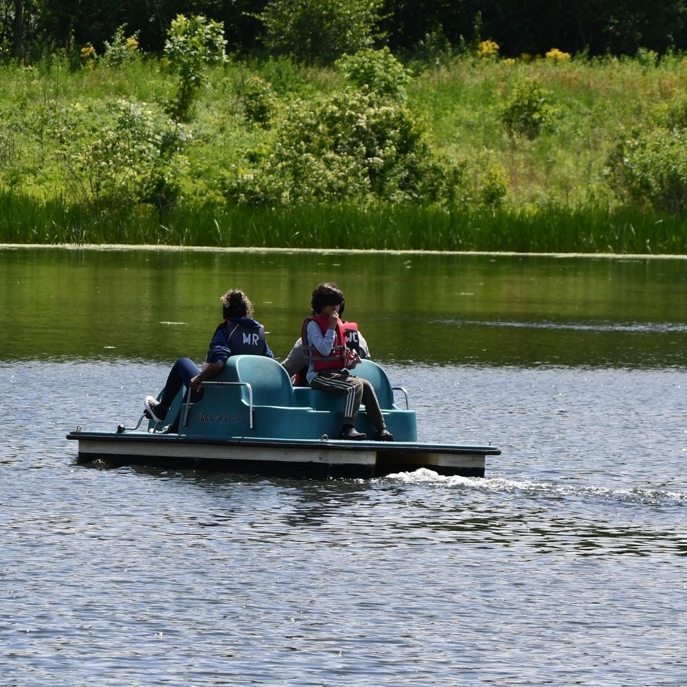 Two people riding a small pedal boat on a calm lake, surrounded by lush green vegetation and trees along the shoreline.