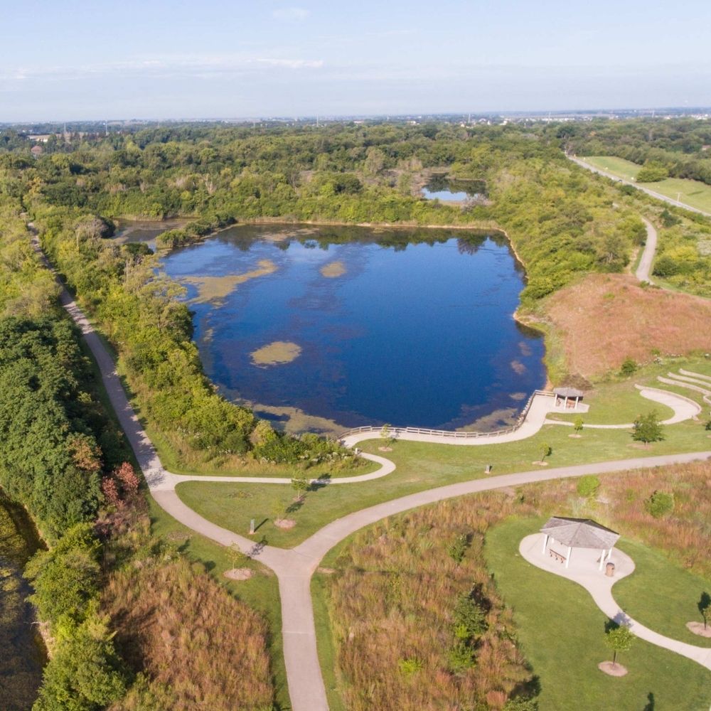 An aerial view shows a large blue lake surrounded by walking and biking trails, grassy prairie areas and dense green trees.