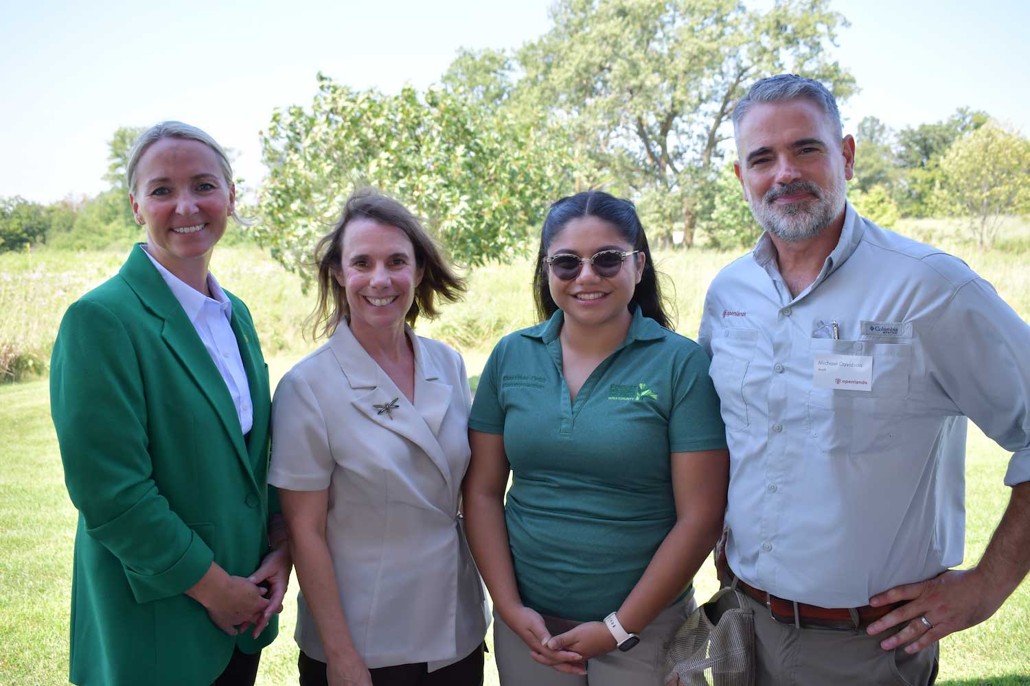 A group of four people standing shoulder to shoulder outdoors with a prairie and trees behind them.