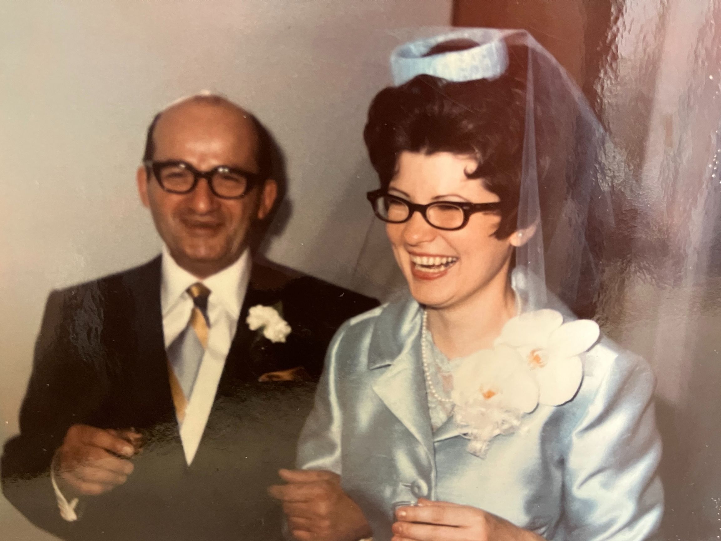 A smiling bride and groom wearing glasses on their wedding day, sharing a joyful moment indoors.