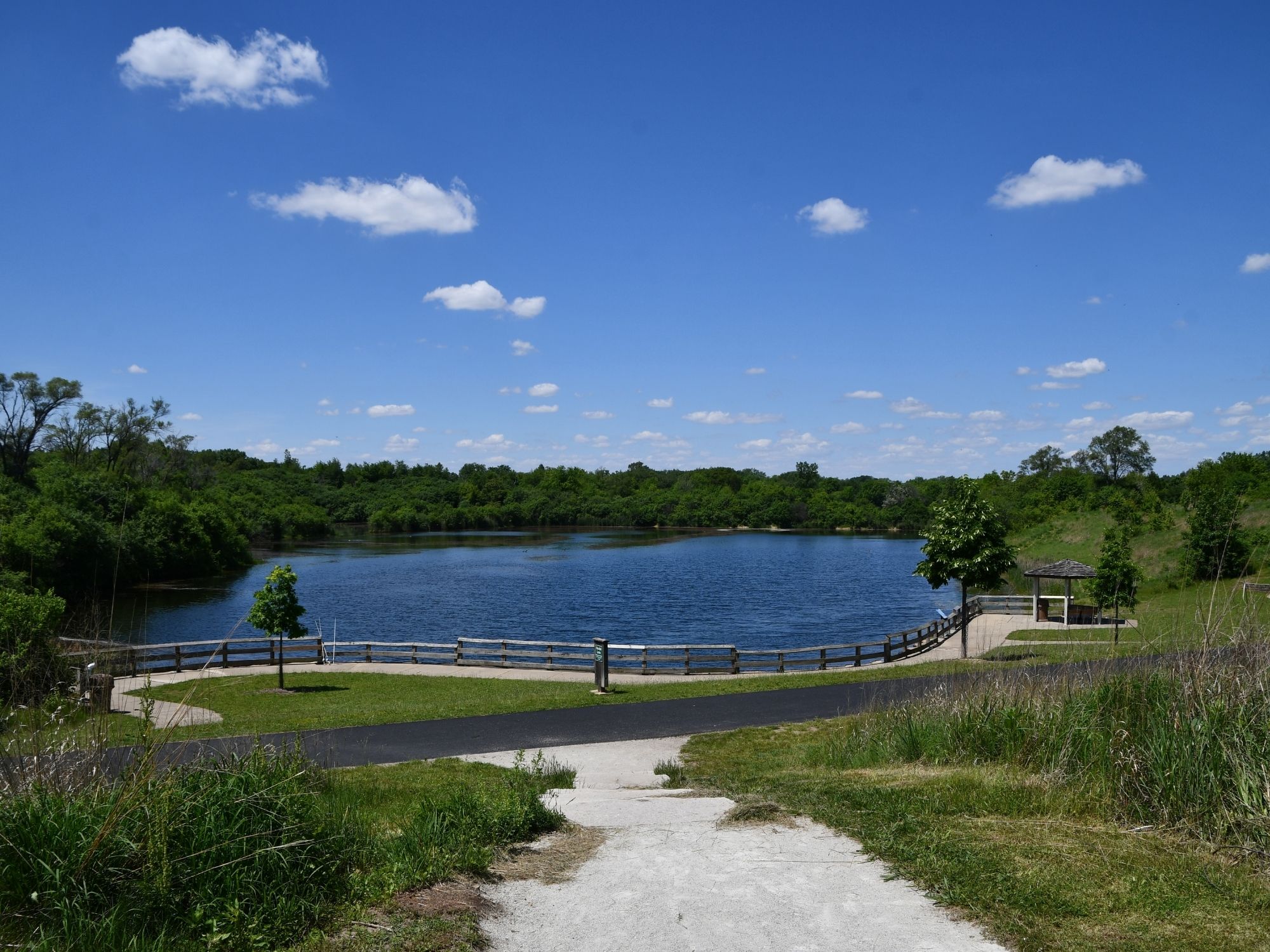 A calm blue lake is surrounded by lush green trees under a bright blue sky dotted with small white clouds.