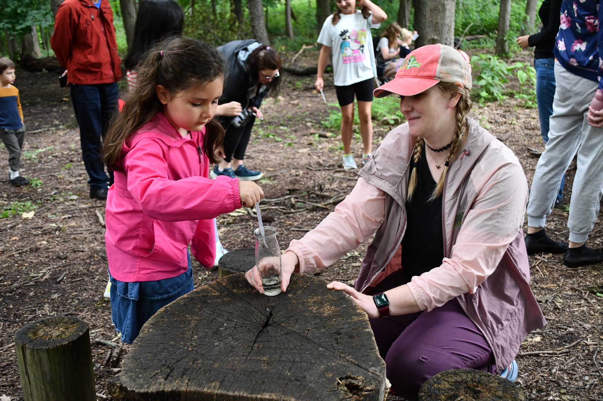 A child putting water in a container held by an adult while standing at a tree stump.