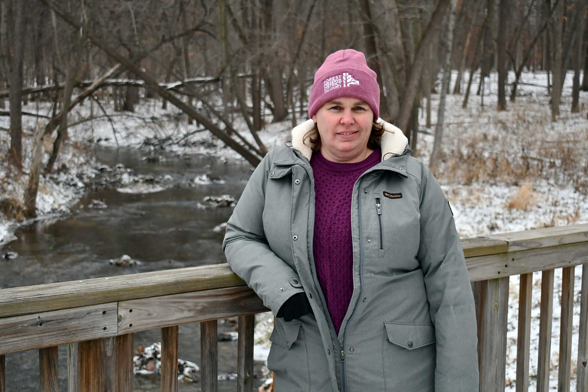 A person stands at a bridge over a creek, wearing a grey coat and pink hat.