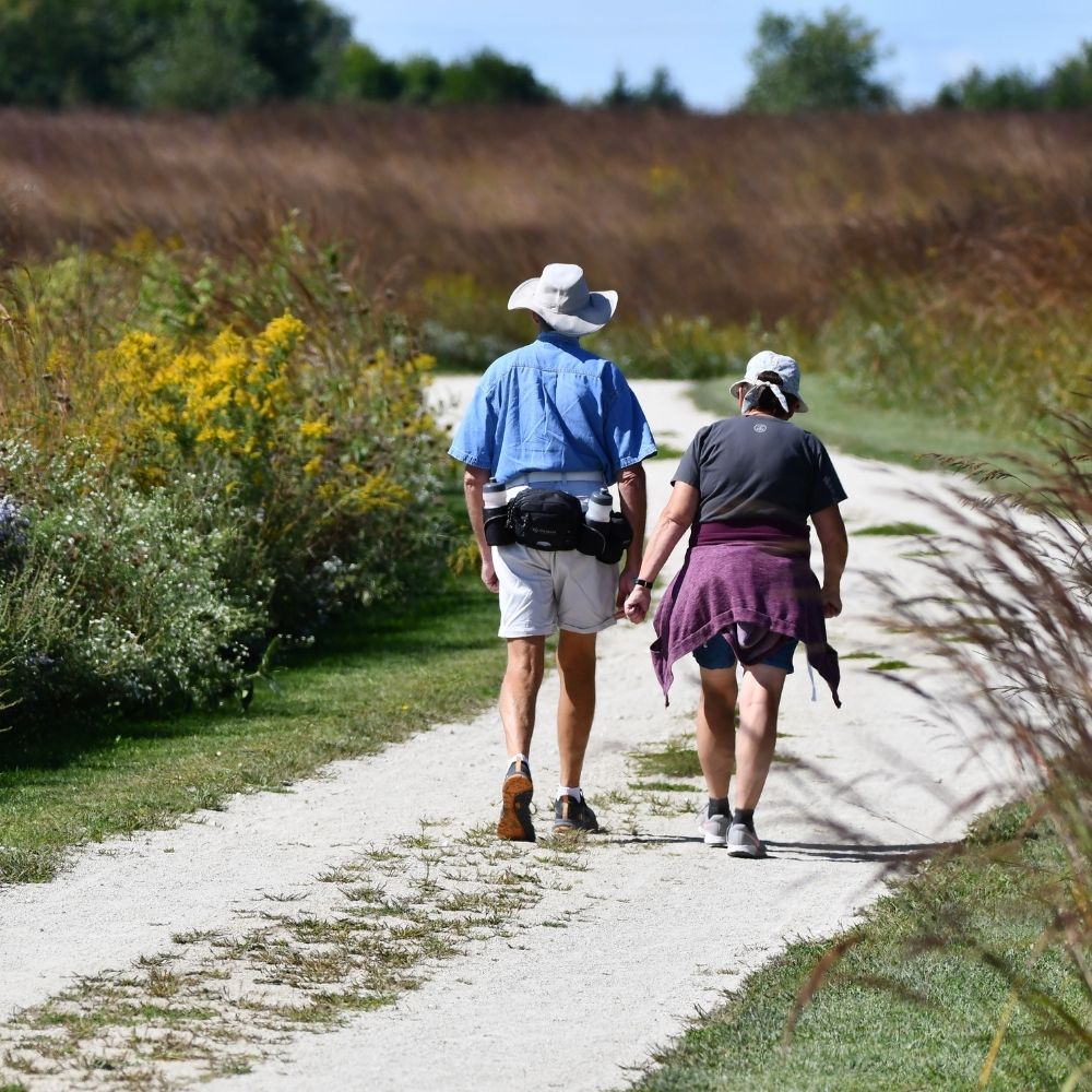 Two people wearing sun hats walk along a gravel trail through a prairie landscape with tall grasses and wildflowers on a sunny day.