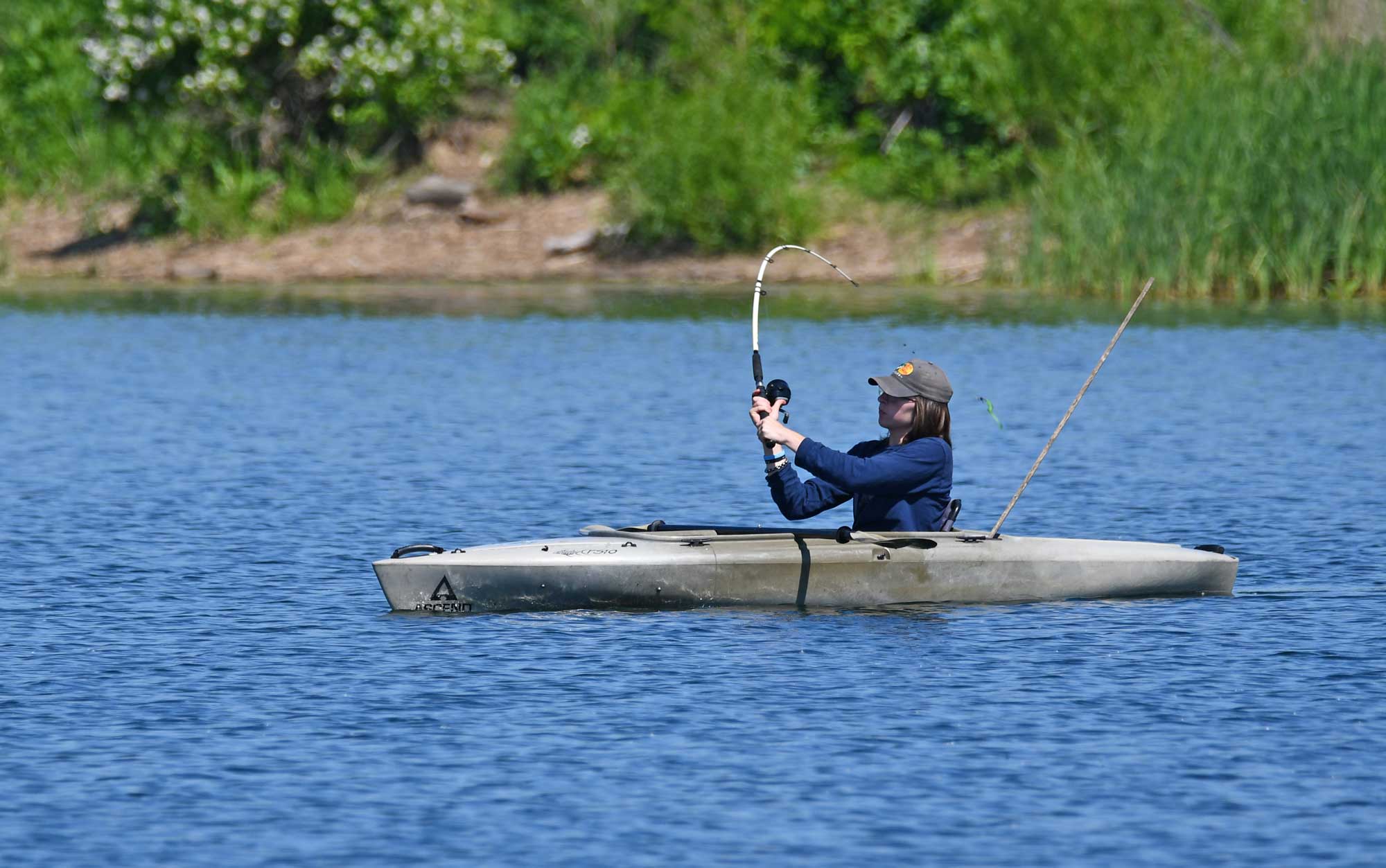 Person fishing in lake while kayaking with vegetation in the background.