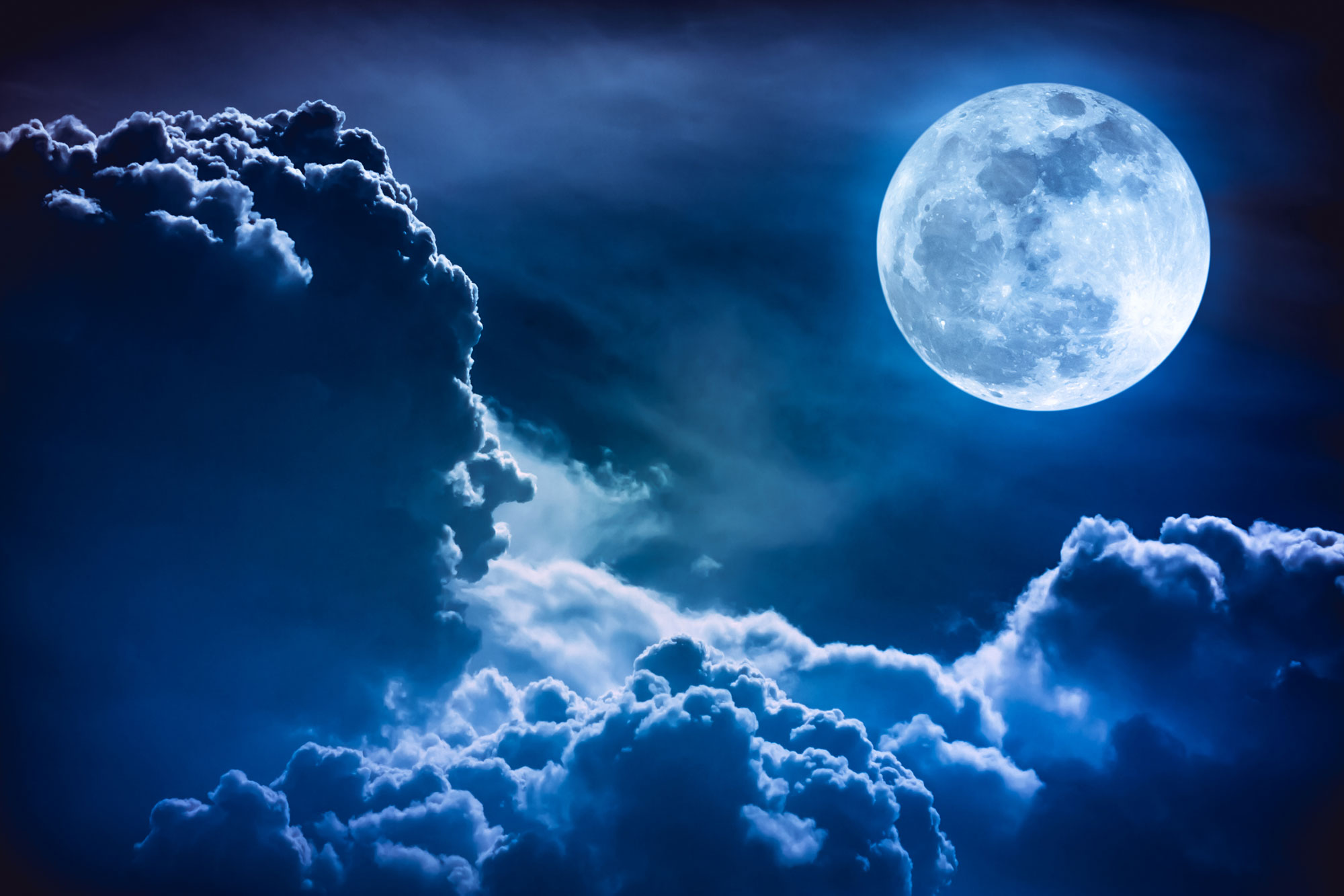 A full moon in a blue hue above clouds in the night sky. 