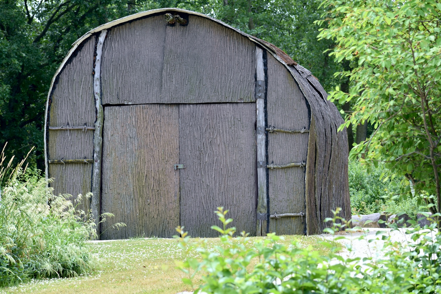 The front of a replica Native American longhouse surrounded by green grass, trees and other vegetation. 
