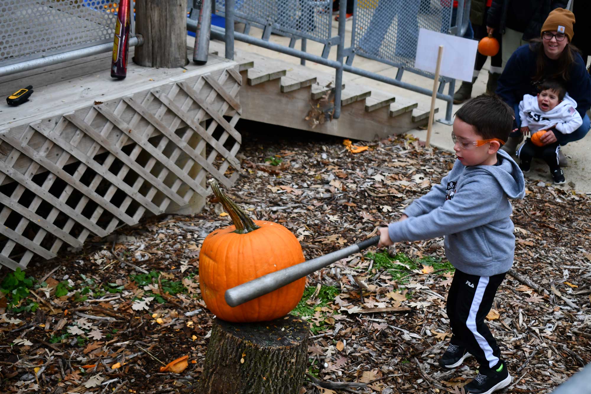 A child using a metal baseball bat to hit a pumpkin on a tree stump. An adult and toddler look on in the background.