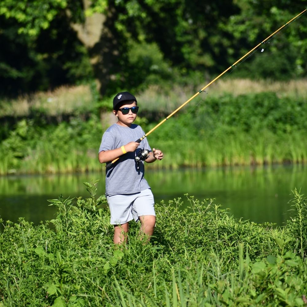 A child wearing sunglasses and a baseball cap fishes along a grassy pond shoreline on a sunny day.