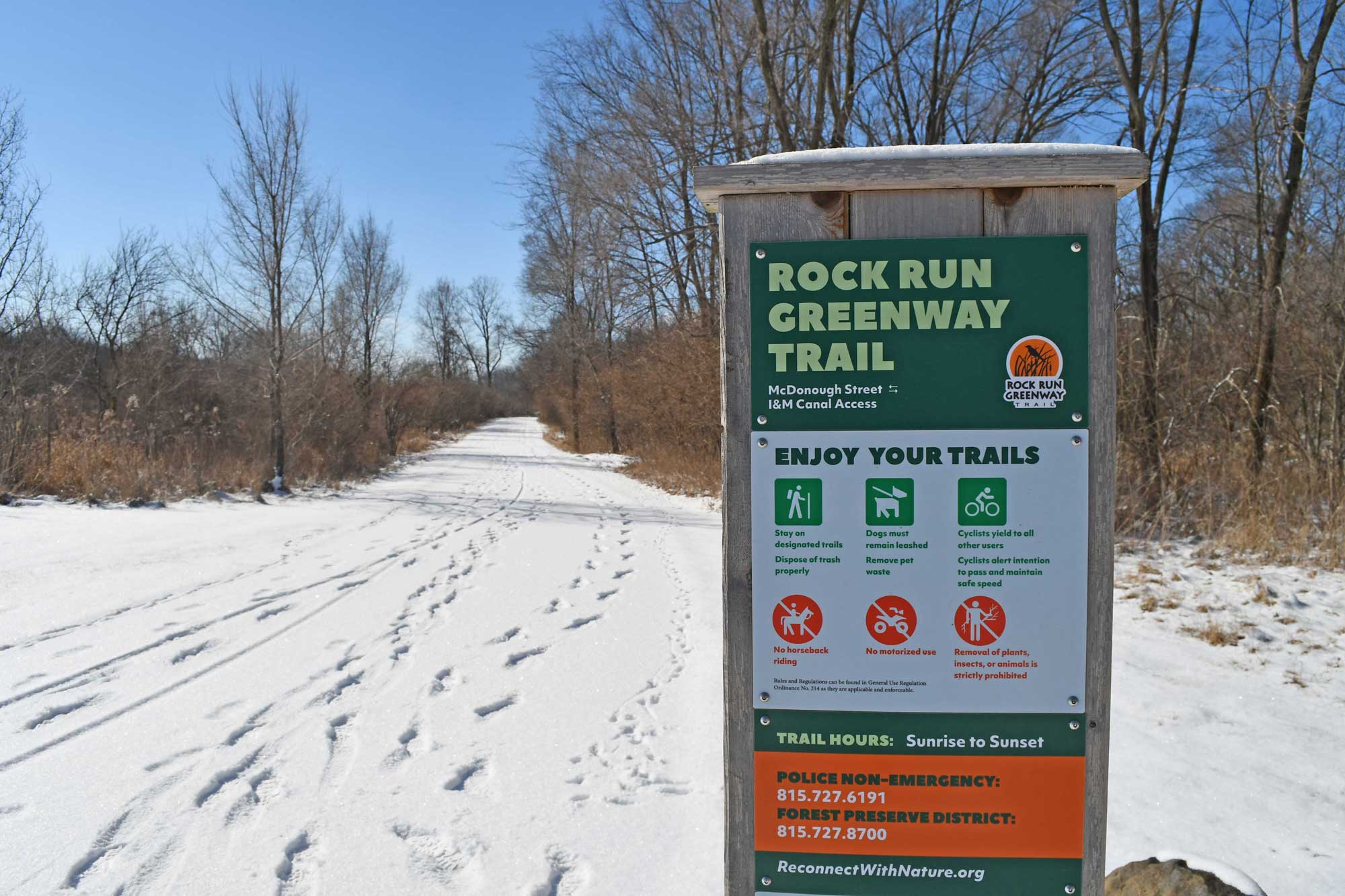 Trail sign with snow covered trail and trees in the background.