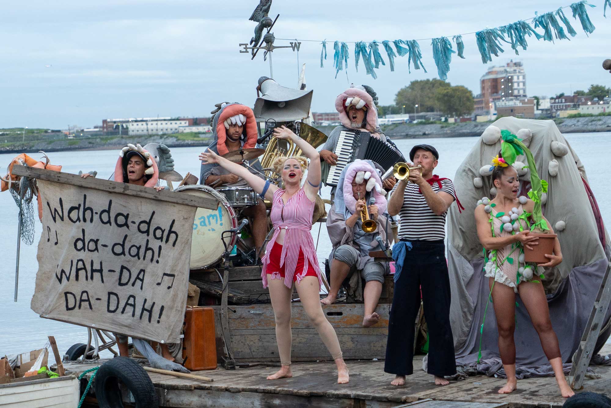 People in circus costumes with instrument on a board with water in the background.