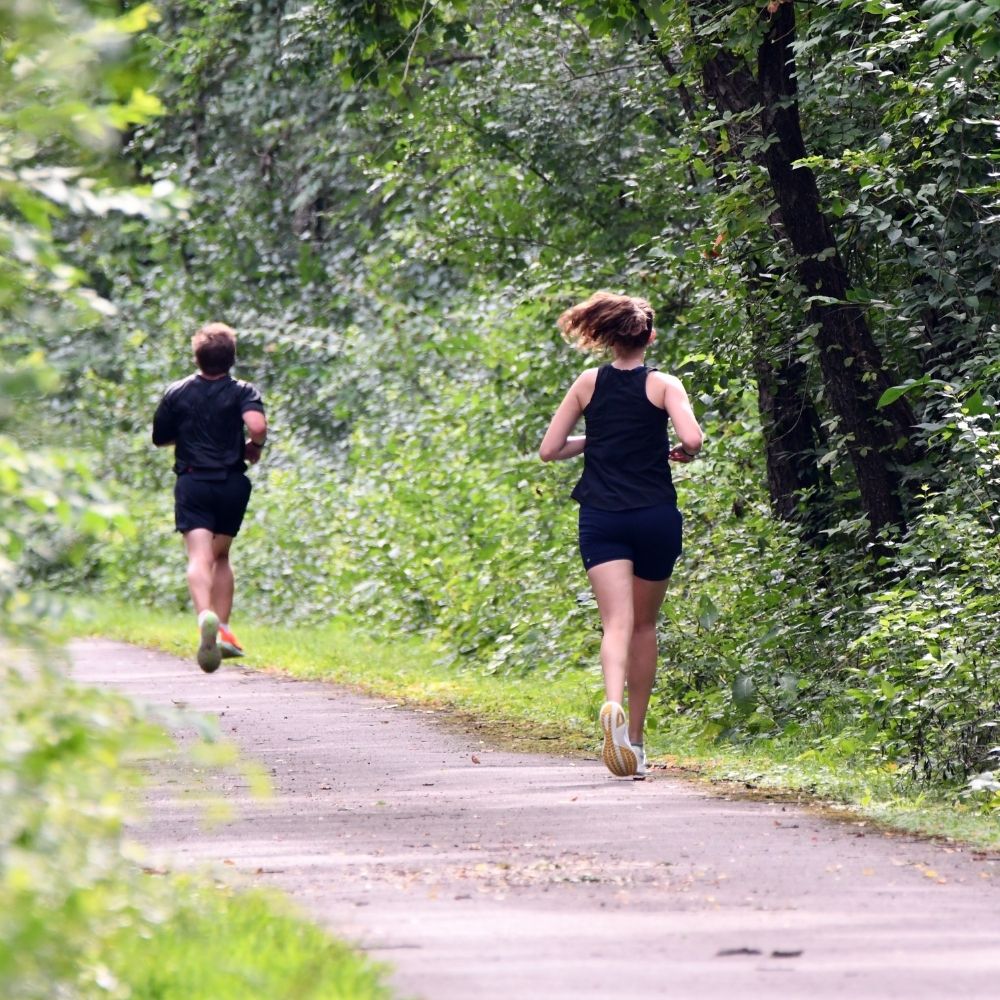 Two people jog along a paved trail surrounded by dense green trees and vegetation.