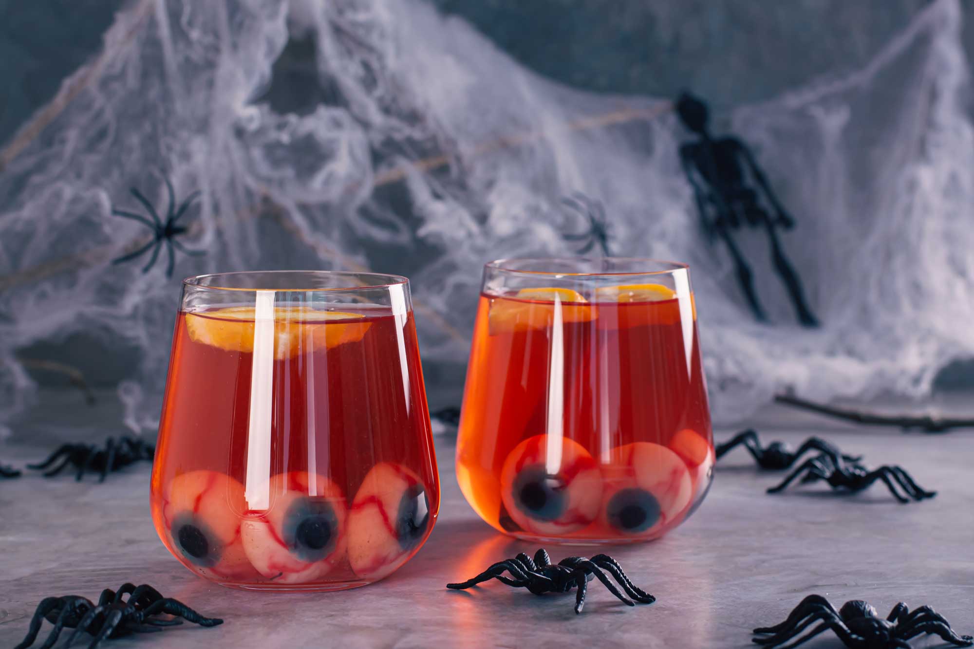 Two cocktail glasses with an orange drink and fake eyeballs set on a table with fake spiders and spiderwebs.