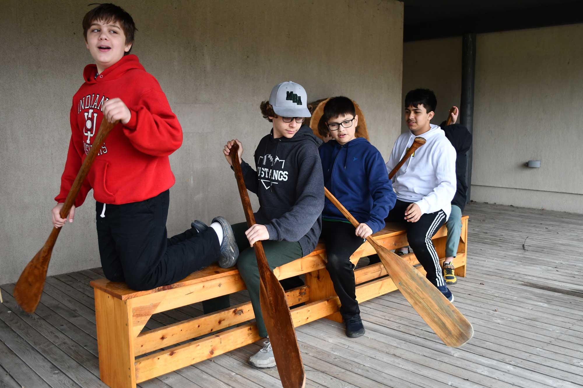 A group of four kids holding oars will sitting on a wooden bench.