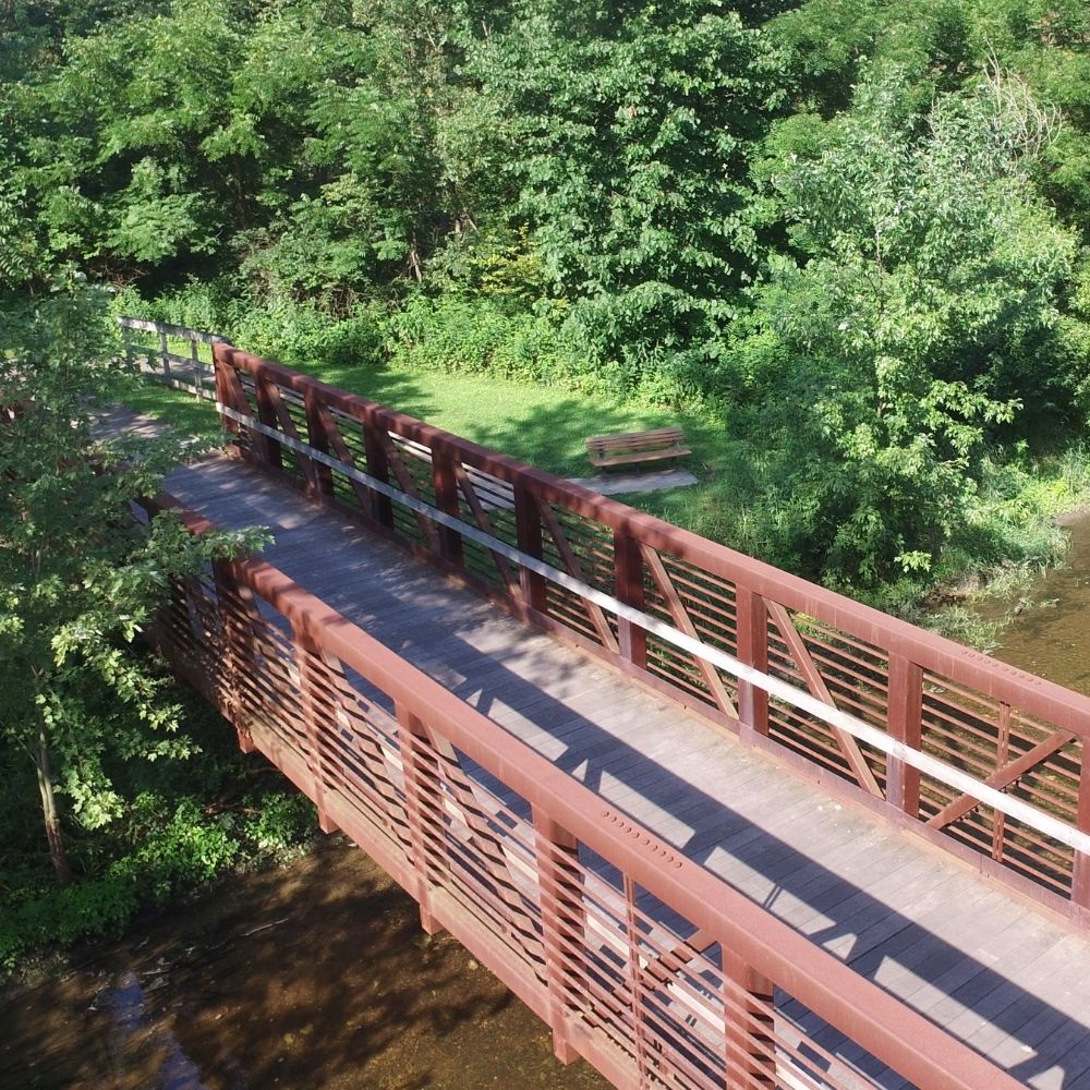 An elevated wooden and metal pedestrian bridge crosses over a shallow stream, surrounded by dense green trees and vegetation, with a bench visible near the trail.