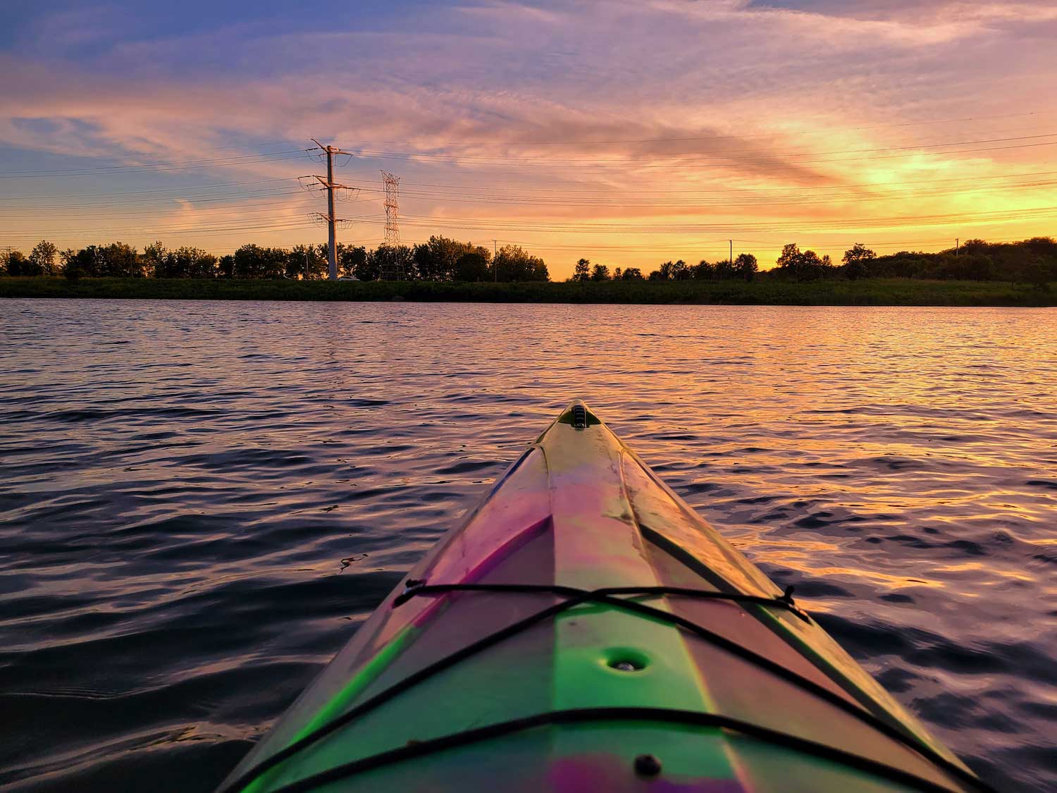 The tip of a colorful kayak on the water with the sky orange from the sunset in the background.