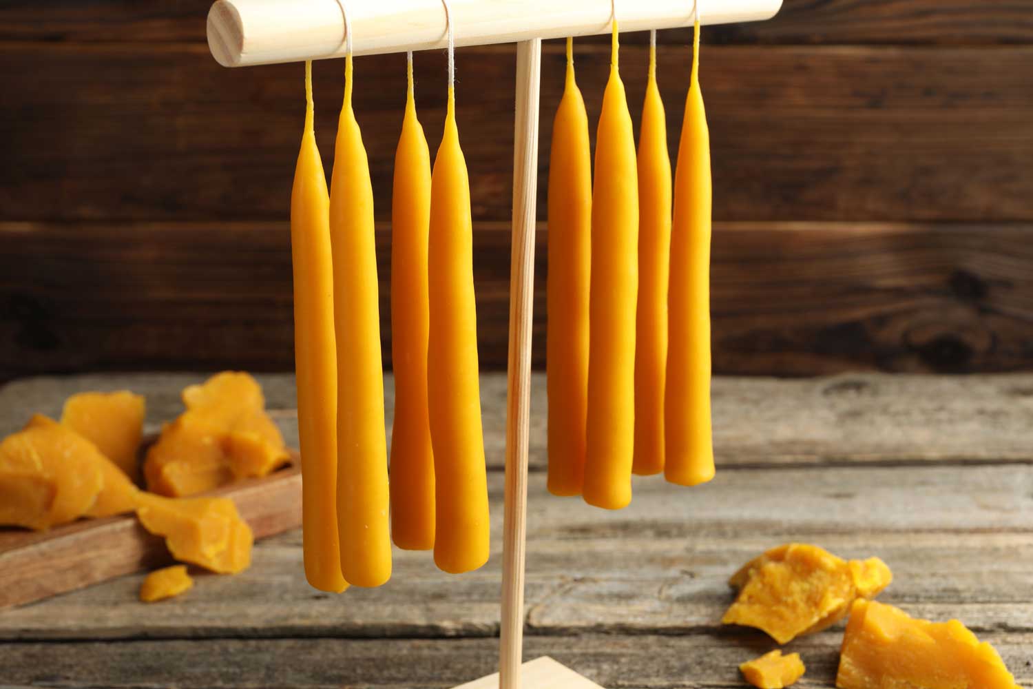 Yellow beeswax candles hanging to dry on a wooden rack with chunks of beeswax laying on a wooden table. 