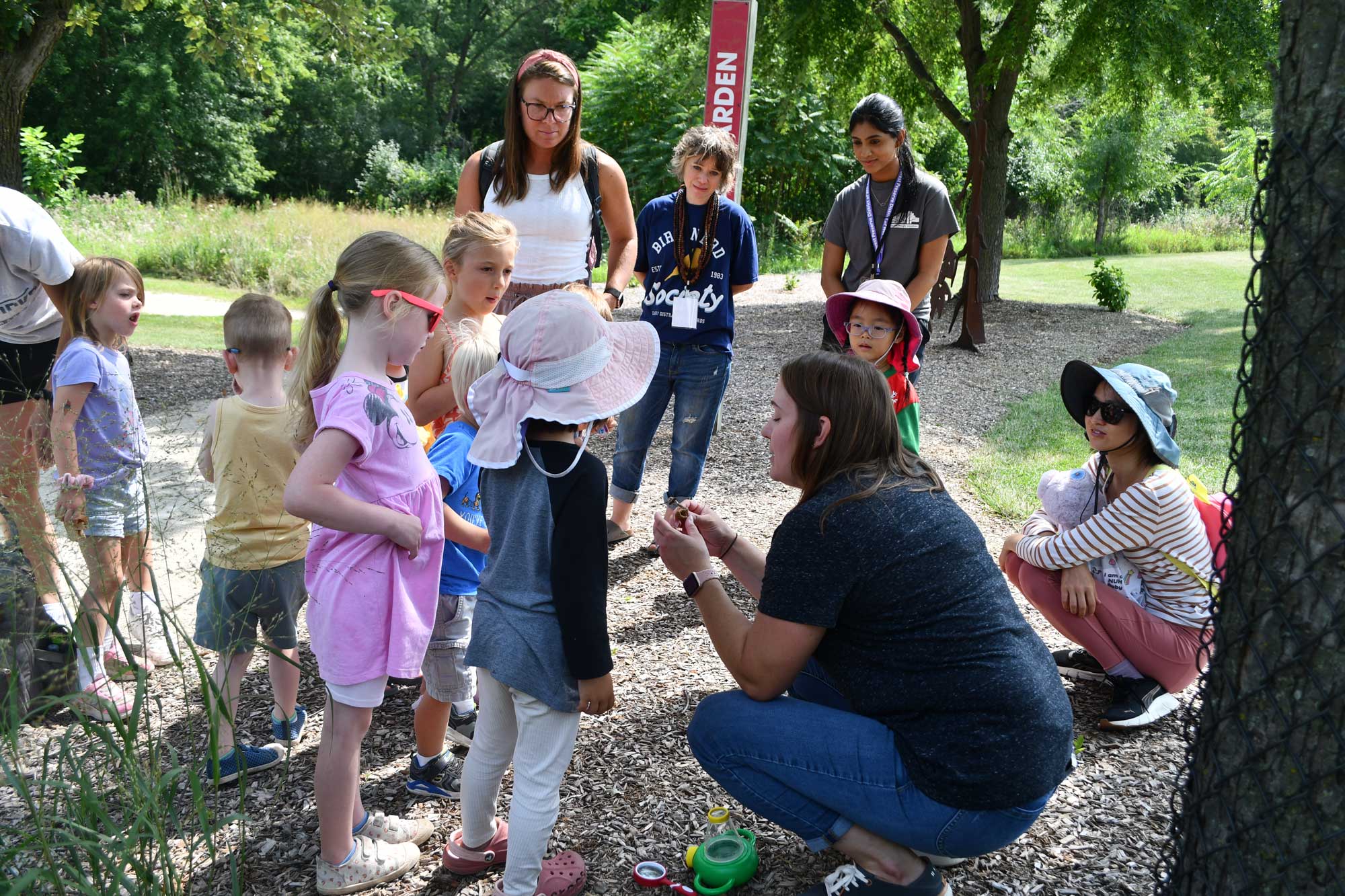 An adult crouched down along an outdoor trail showing a group of kids something while adults look on in the background.