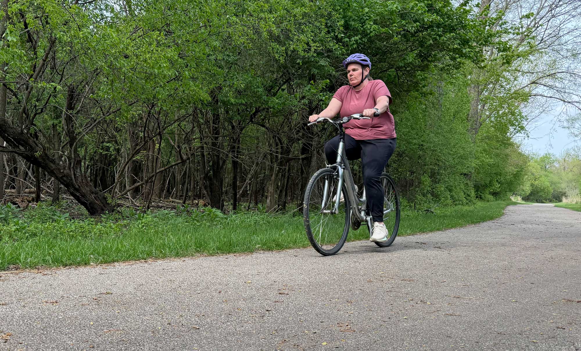 A person wearing a maroon shirt and black pants rides a bike on a paved trail that runs through woodlands. 