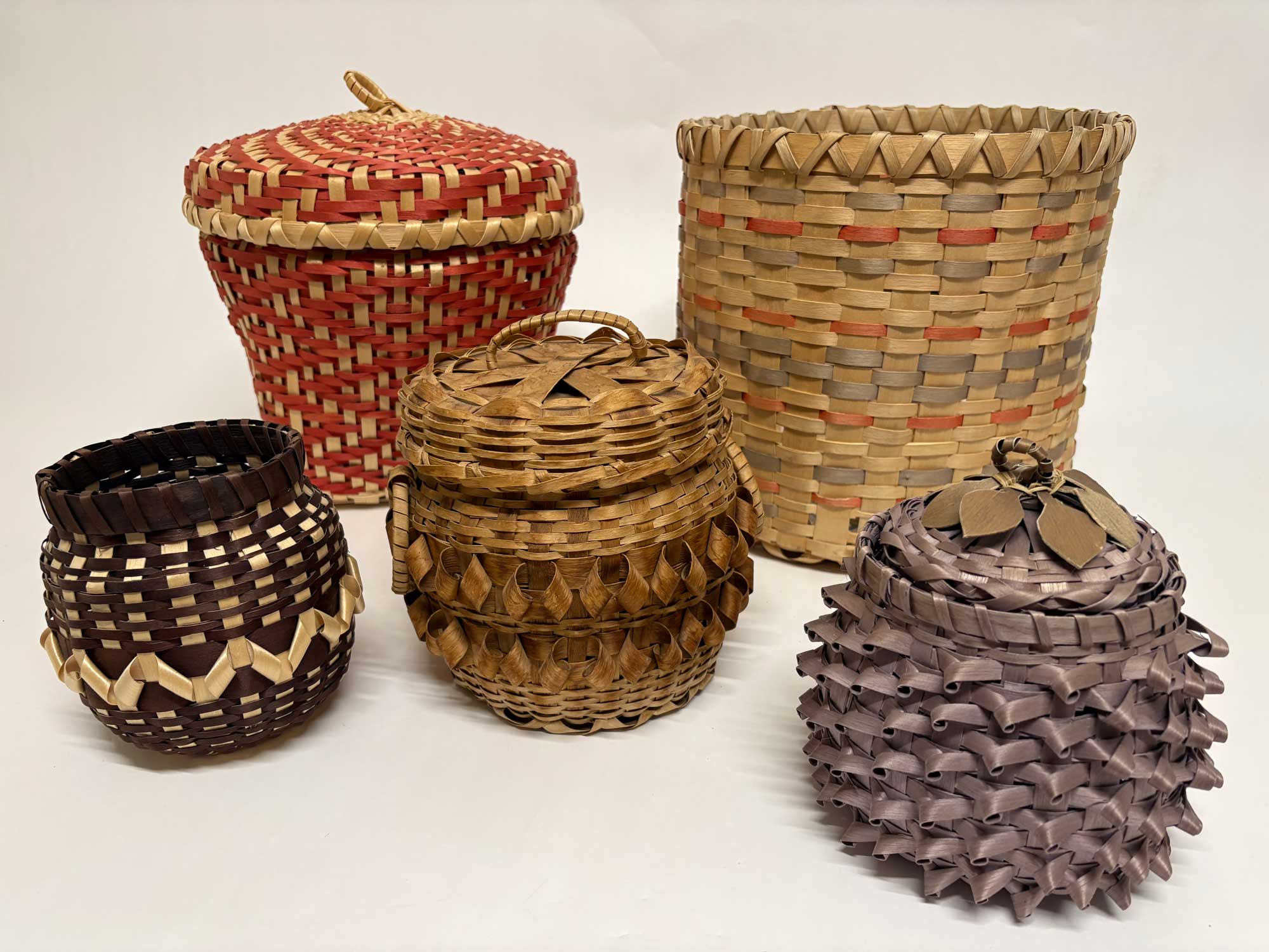 A collection of five woven Potawatomi baskets in shades of brown, red and blue resting on a white surface with a white background.  