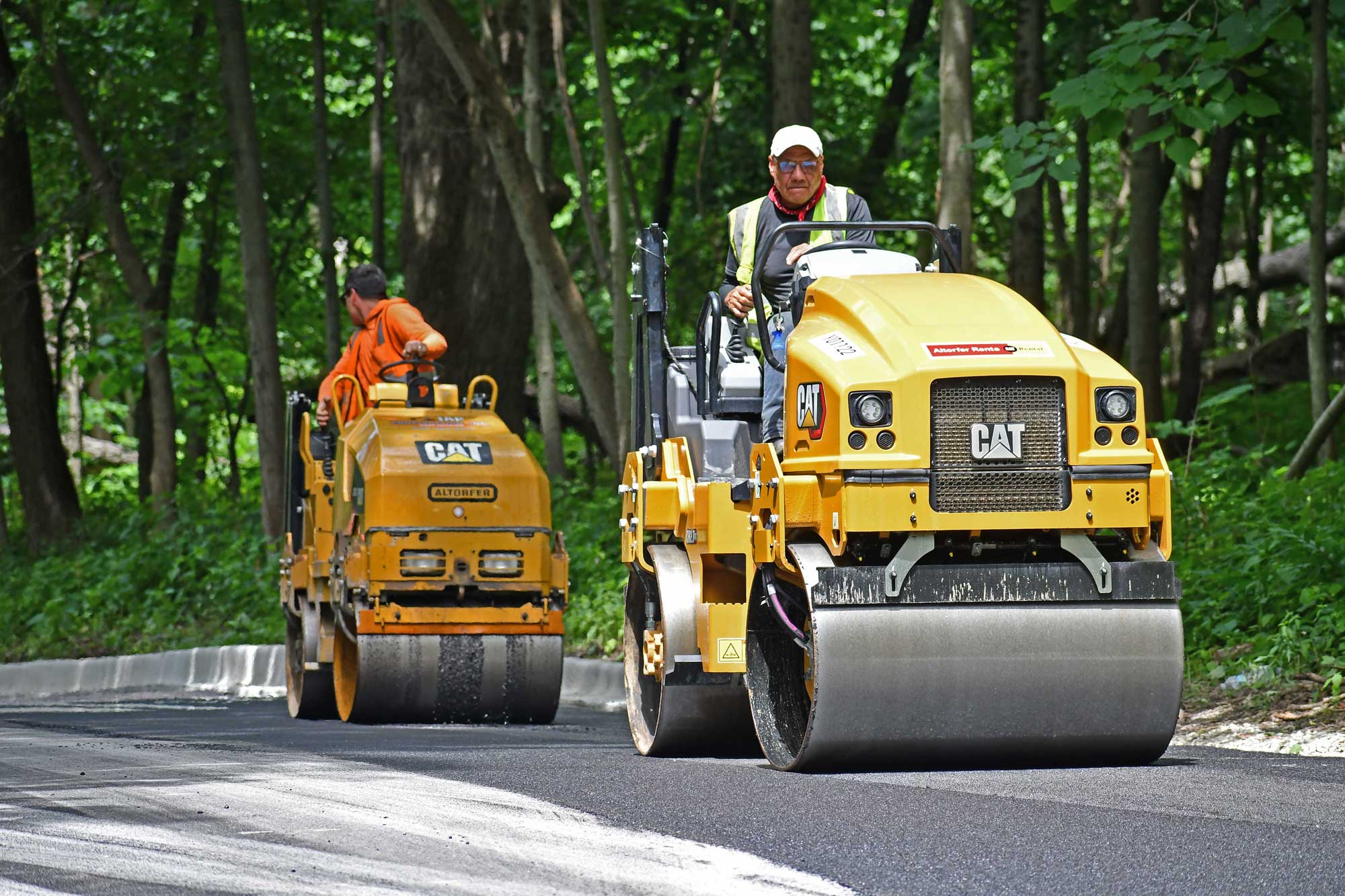 Two road rollers moving along a newly paved ashphalt road, with trees visible in the background. 