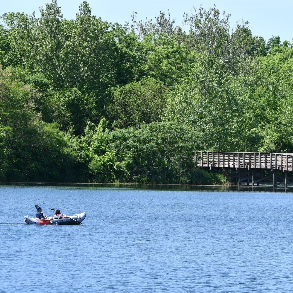 Two people paddle an inflatable kayak across a calm lake on a sunny day, with a wooden boardwalk and dense green trees along the shoreline in the background.