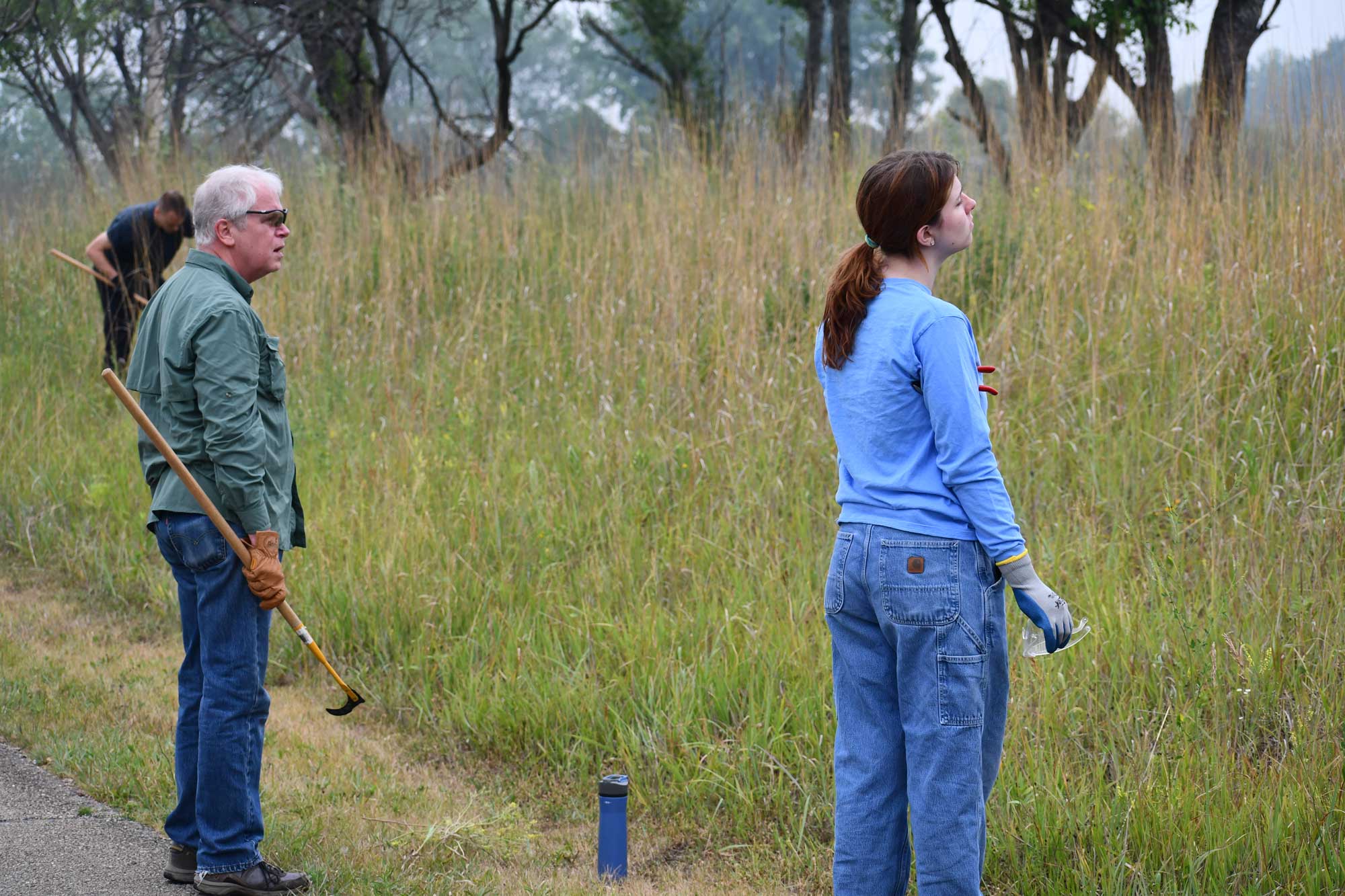 Two people holding landscape tools and protective gear standing at the edge of a grassy area looking out into the field.