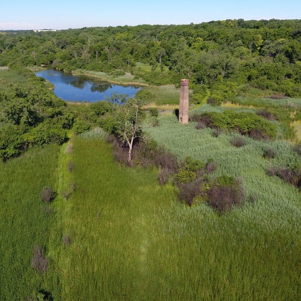 An aerial view of a green wetland landscape with a winding river, dense trees, and tall grasses surrounding a lone brick chimney rising from the marsh.