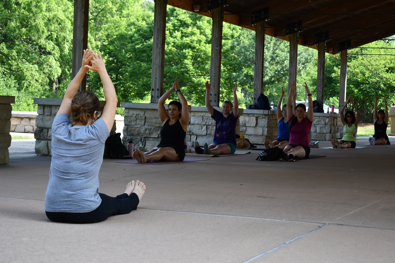 A person leading a group in yoga. The group is sitting on the ground with legs extended in front of them and arms above their heads. 