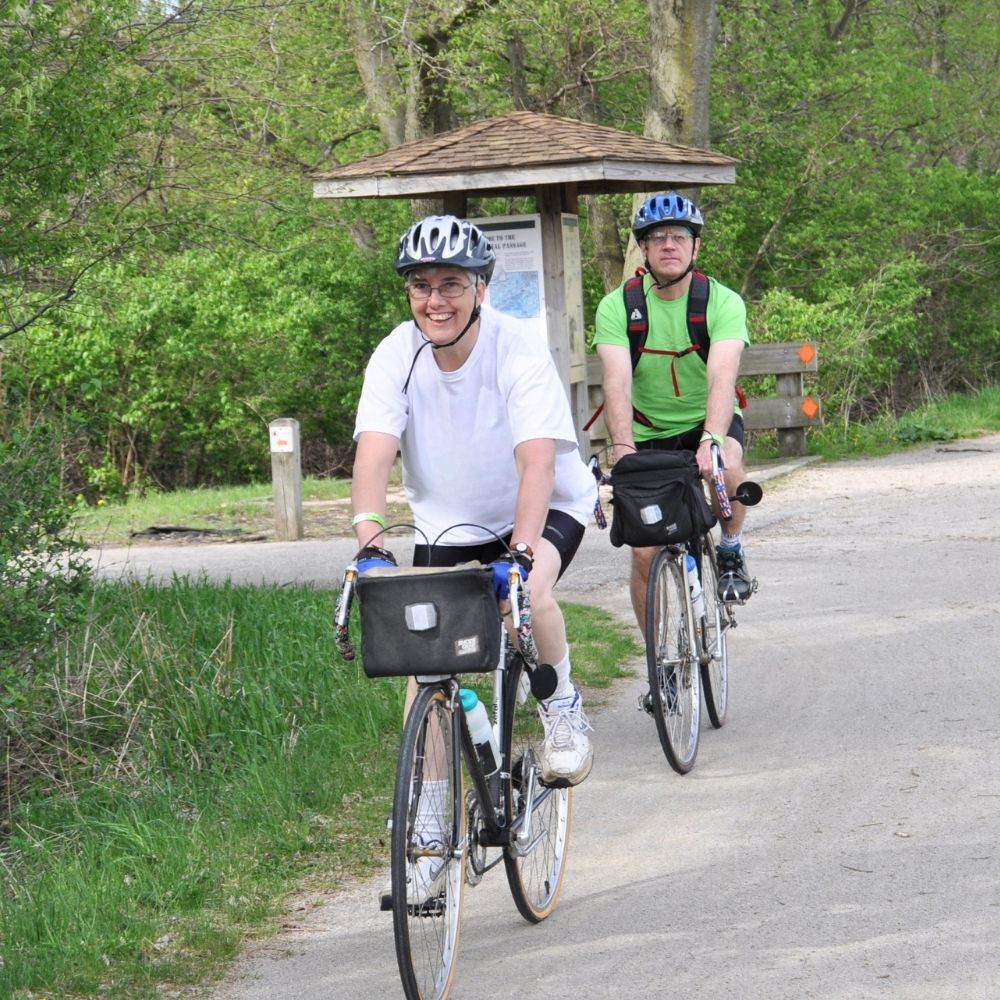 Two people wearing bicycle helmets ride bikes along a paved trail through a wooded park, passing a trail information sign on a sunny day.