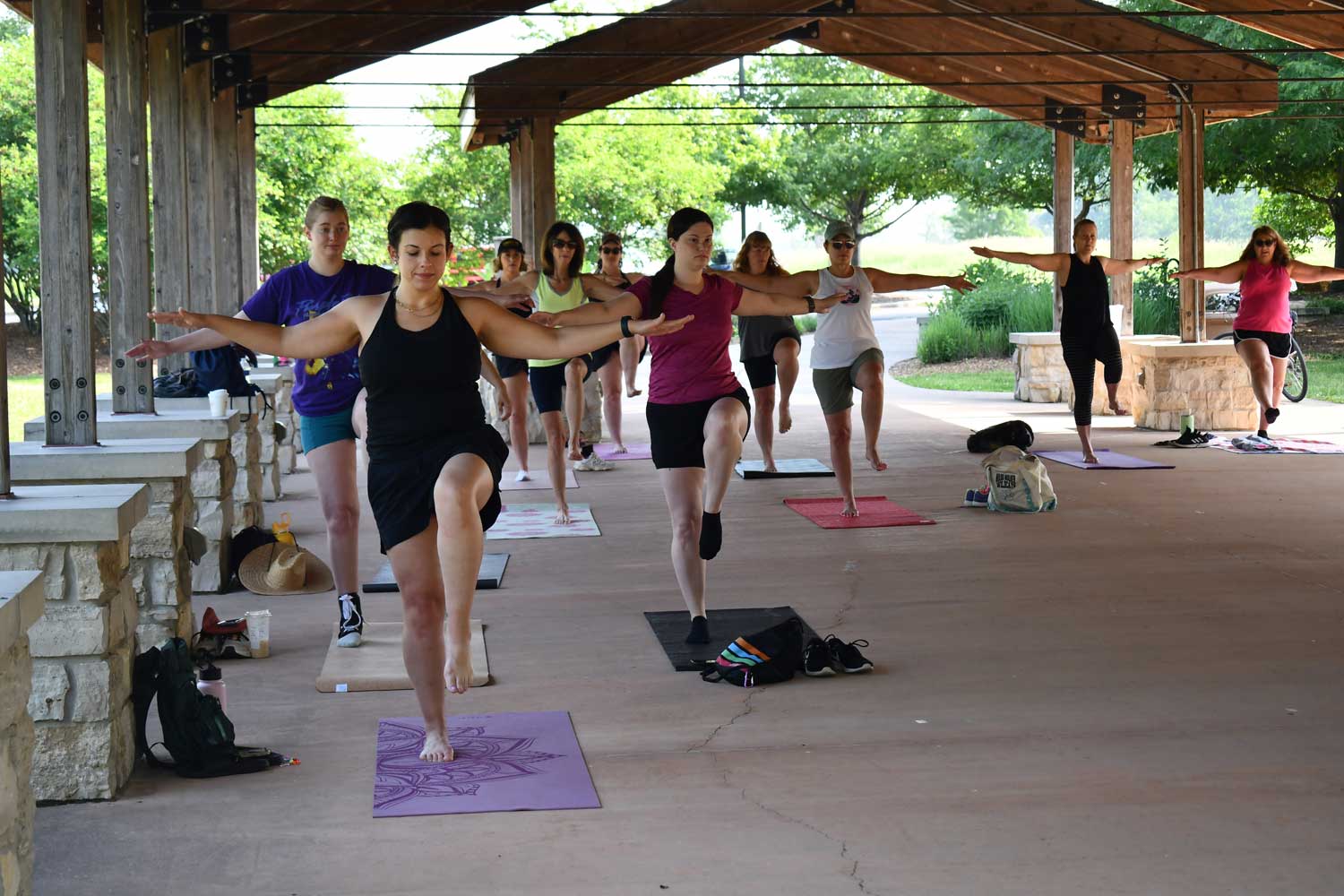 A group of people standing on yoga mats under an outdoor shelter. The group is standing on one foot with their arms stretched out at their sides.