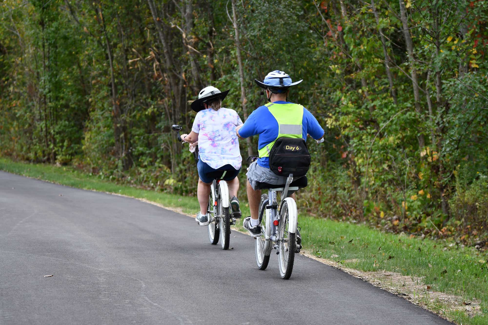 Two people biking on a paved trail lined by grass and vegetation.