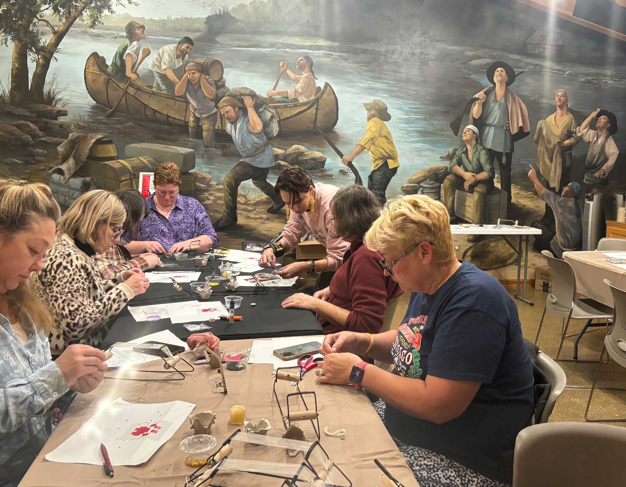A group of adults sitting at a long table bent over looking at beading projects. Beading supplies are laying on the table. 