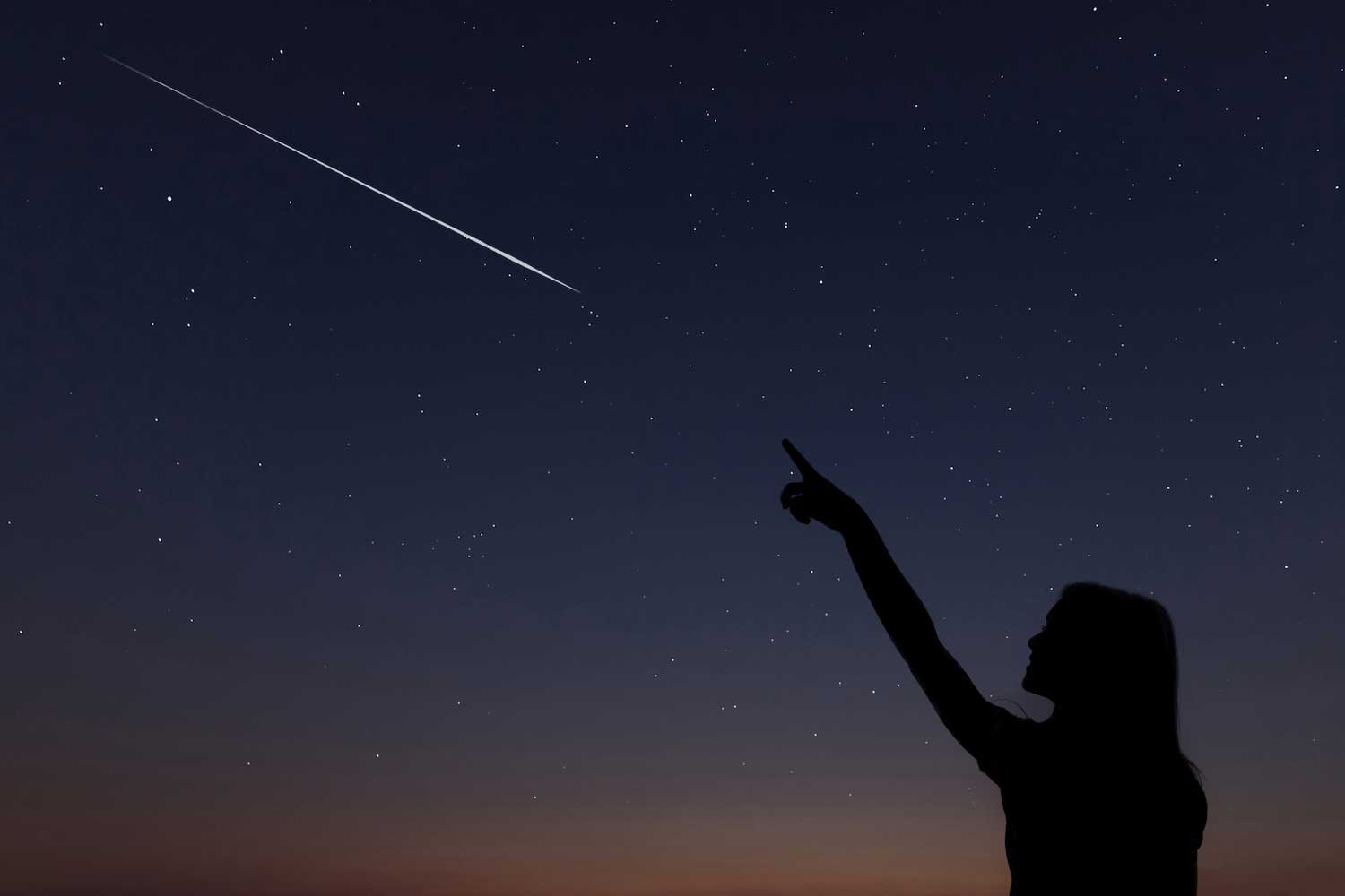 The silhouette of a person standing in the dark pointing at a shooting star streaking across the star-filled sky.