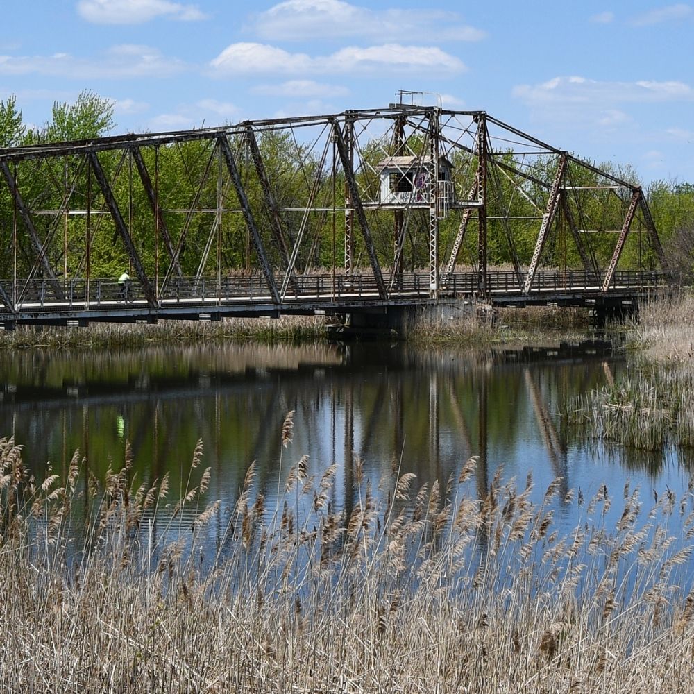 Historic truss bridge over calm water with spring trees and tall grasses reflected in the surface beneath a blue sky with scattered clouds.