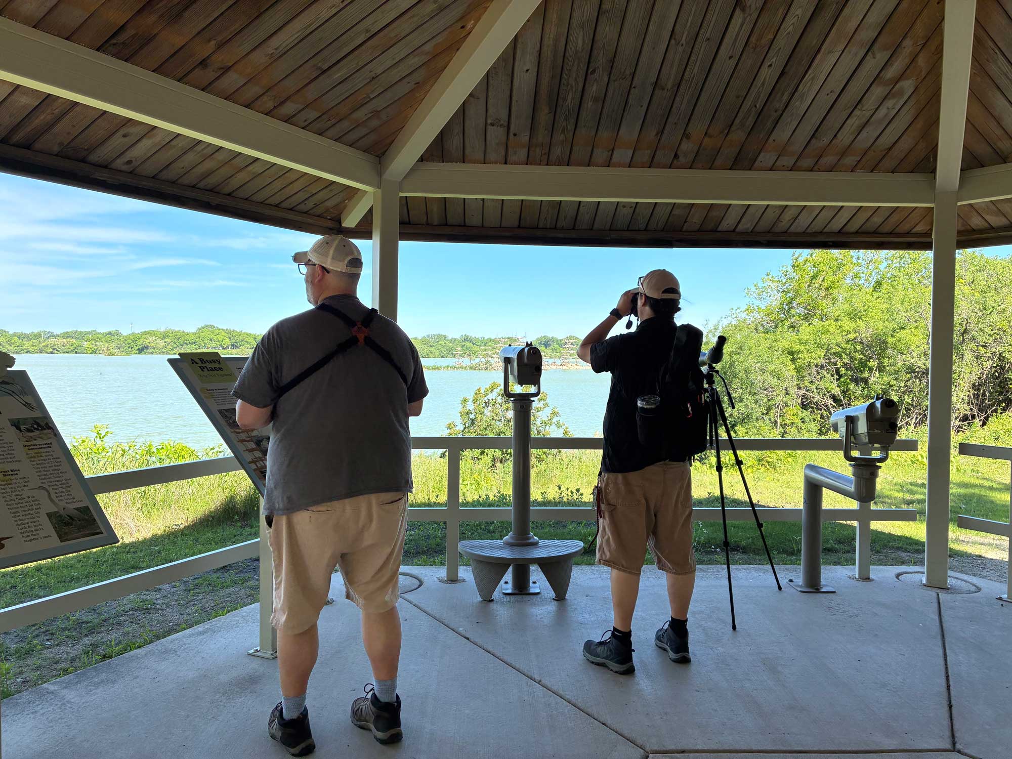 Two people standing under a shelter looking out at a lake. One person is looking through binoculars. Spotting scopes and interpretive signage is in front of them.