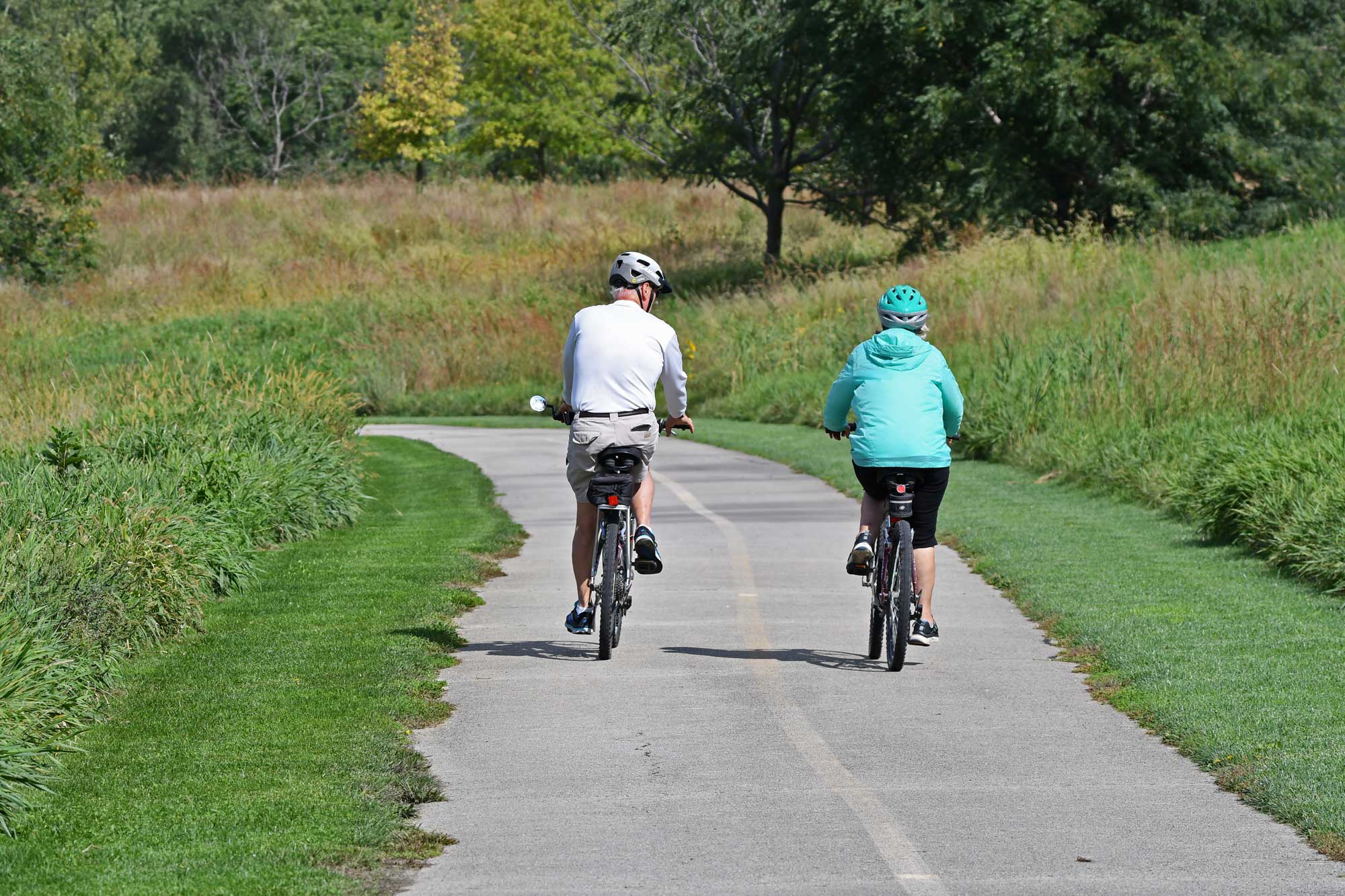 People riding bikes on a paved trail with vegetation and trees in the background.