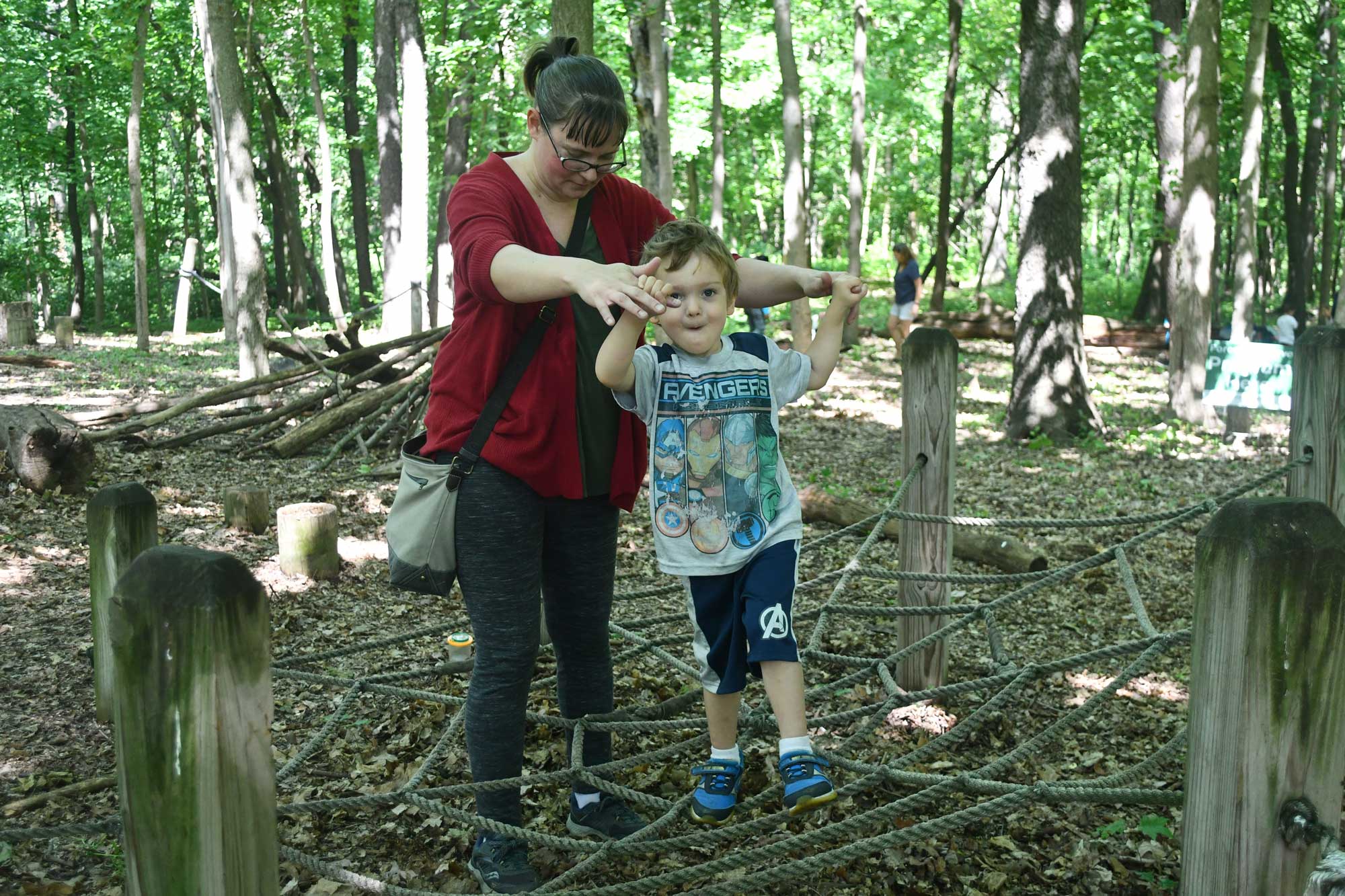 An adult helps a child walk across a rope play structure shaped like a spiderweb.