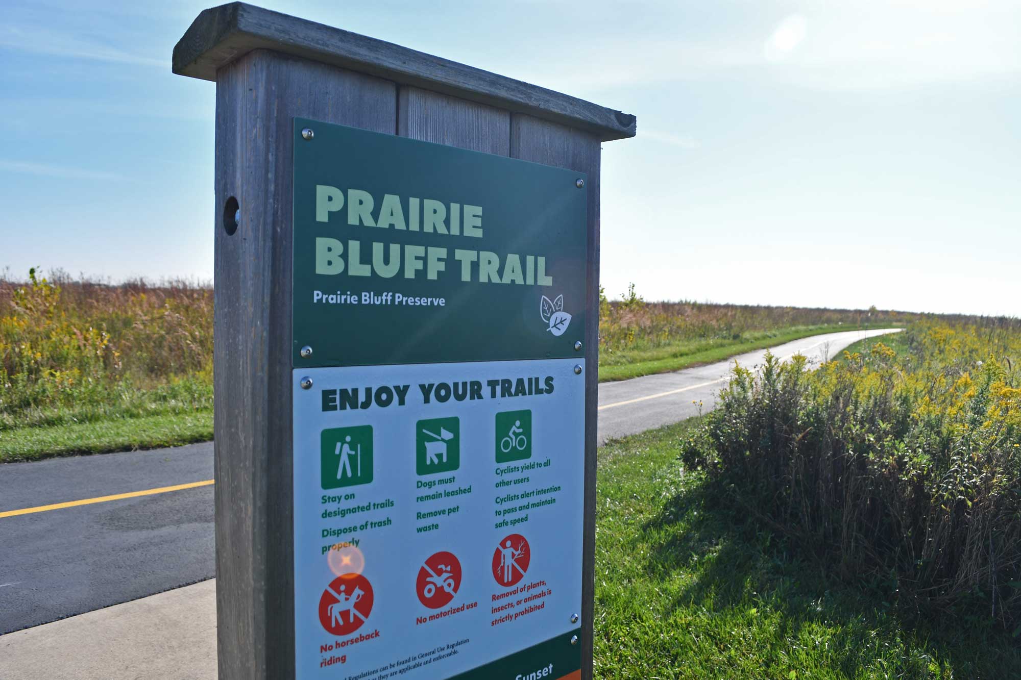 Trail sign with paved trail and vegetation in the background.